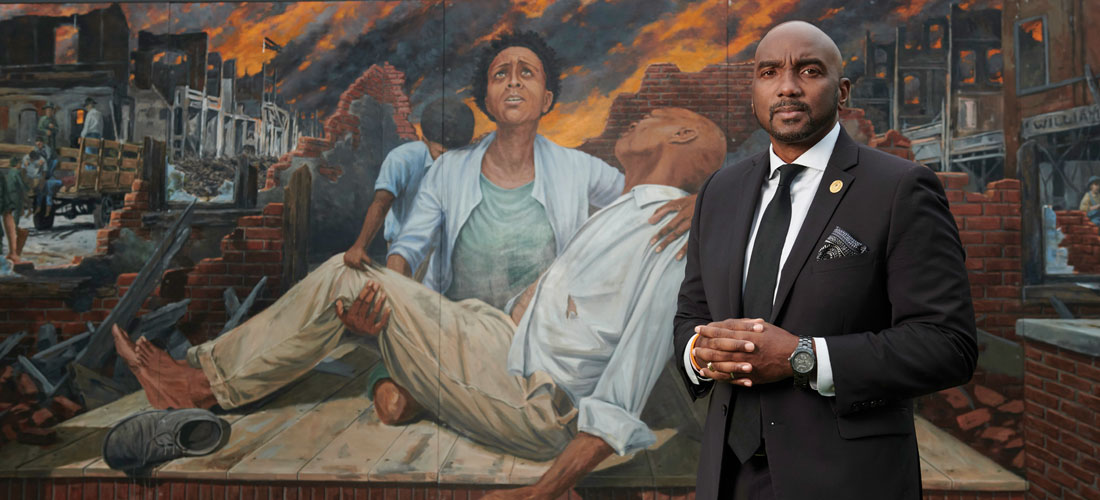 Black man standing in front of a mural where a Black woman cradles an injured or dead young man, as in the Pieta stature of the Virgin Mary cradling a dead Jesus.