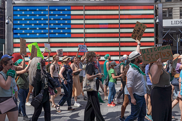 Protestors march past a large US flag mural holding protest signs advocating for abortion rights