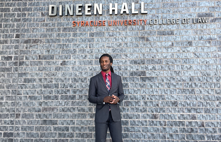 Kendall Anderson stands in front of a wall with a sign reading Dineen Hall, Syracuse University College of Law