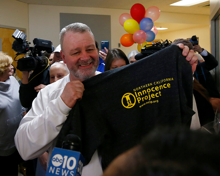 A smiling man holds a t-shirt with the logo of the Northern California Innocence Project as he is surrounded by news cameras with ballons in the background