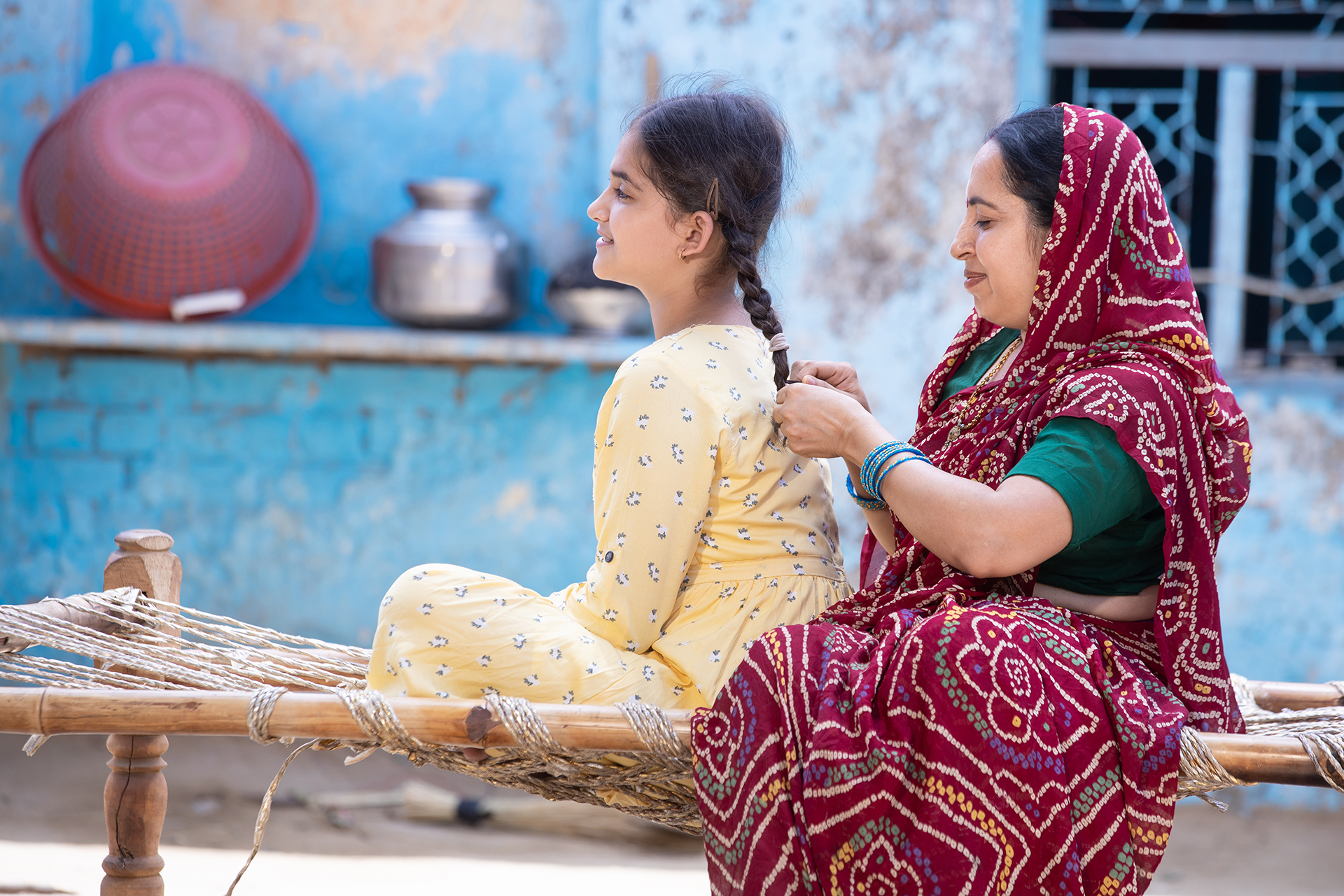 An Indian mother sits behind her daughter, braiding her hair. Both are smiling.