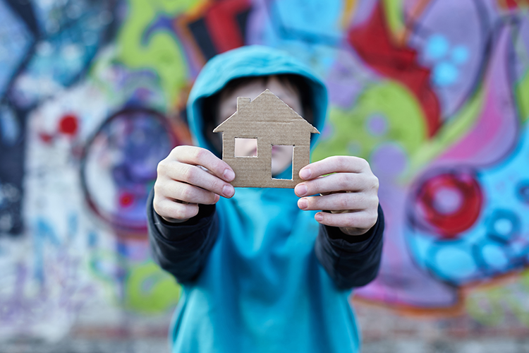 Youth holding up a cutout of a home in front of a wall of graffiti