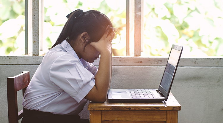 Sad little girl with her head in her hands sits at a desk looking at her computer.
